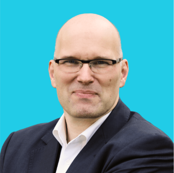 Um homem careca com óculos, usando uma camisa branca e um blazer escuro, posa contra um fundo azul sólido.