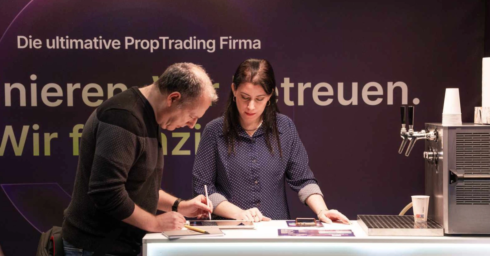 Un homme et une femme signent des documents à un stand avec un robinet et des gobelets à proximité.