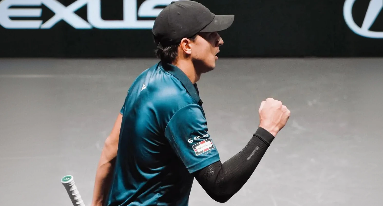 Un giocatore di tennis con maglia verde acqua e cappellino nero alza il pugno, con il logo SabioTrade visibile.