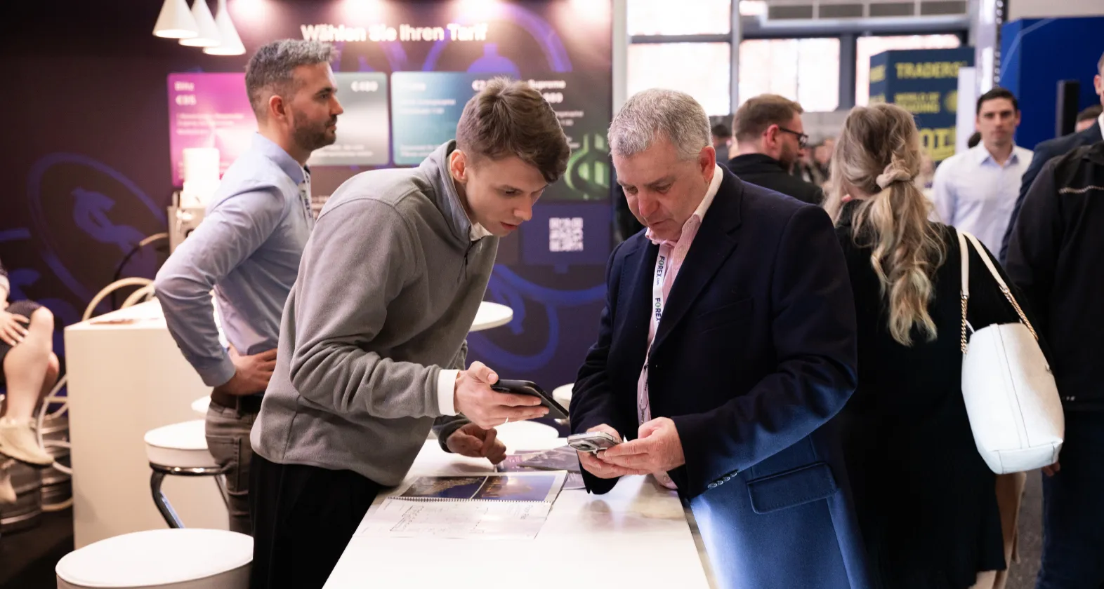 Due uomini esaminano un display SabioTrade a una fiera, con diversi partecipanti nelle vicinanze.